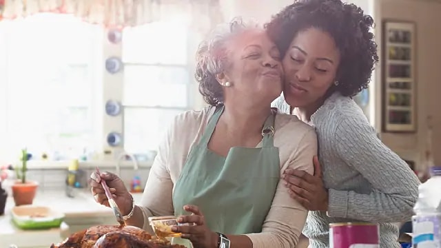 A woman is kissing her mother on the cheeks while her mother is making turkey